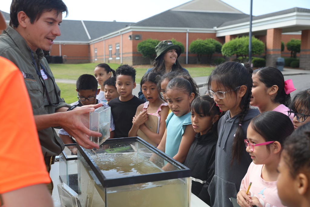 A group of elementary-age students gather outdoors around a table during a hands-on science activity. An instructor demonstrates something inside a large water-filled tank while the children watch closely with curiosity and excitement. Several students hold clipboards and pencils, and a school building is visible in the background on a sunny day.