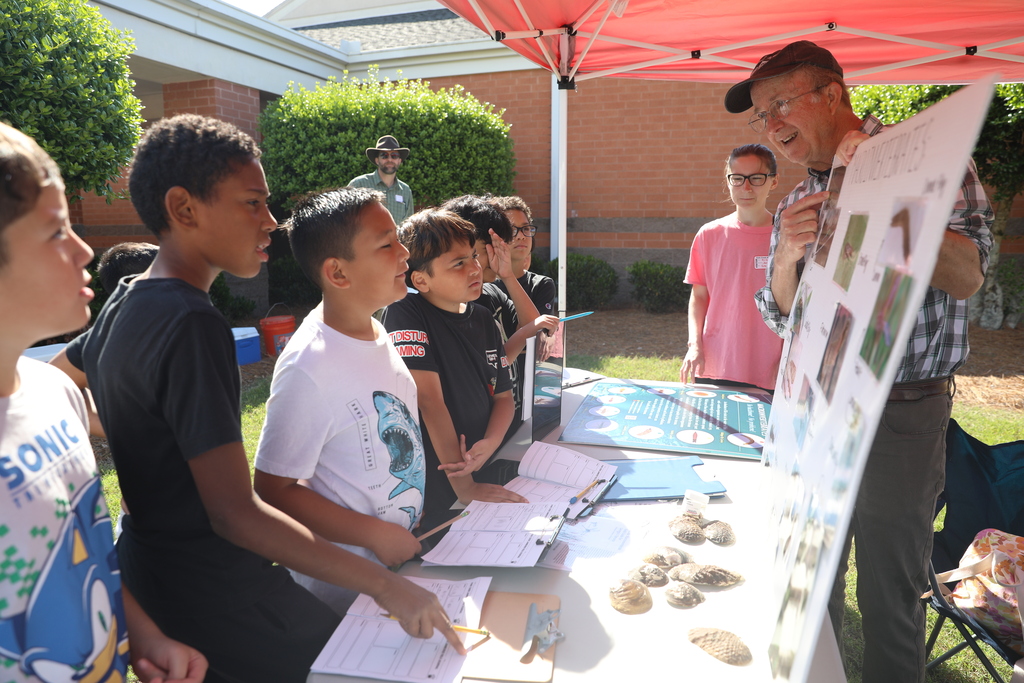 A group of elementary-age students stand around an outdoor learning station under a red canopy tent while listening to a presenter explain a display board with images and information. The children hold clipboards and worksheets as they participate in the hands-on science activity. Seashells and educational materials are spread across the table, and school buildings and greenery are visible in the sunny background.
