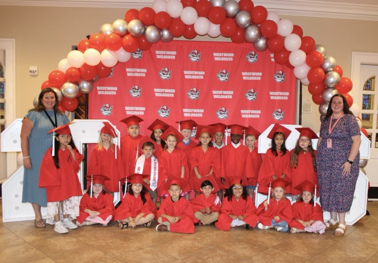 Mrs. Smith’s abd Mrs Atkinson’s class wearing their caps and gowns for graduation standing in front of the ballon arch and Westwood backdrop 
