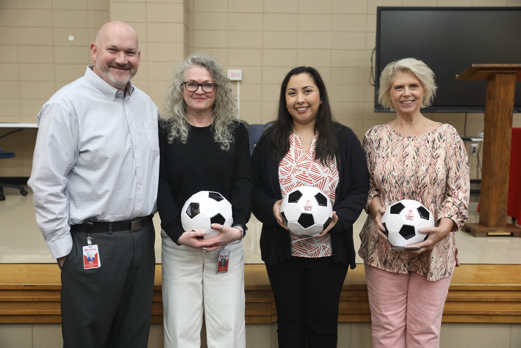 Four Dalton Public Schools staff members stand together indoors during a recognition event, smiling while holding black-and-white soccer-themed “Game Ball” awards. A man in a light dress shirt stands beside three women dressed in professional attire. The group poses in front of a stage area with a podium and large screen in the background.