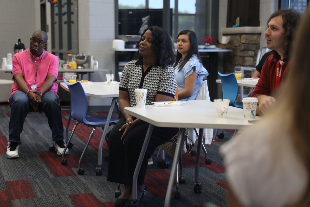 Several staff members sit at tables during a professional development or meeting session inside a modern school library or media center. Attendees listen attentively to a speaker off camera while coffee cups, breakfast items, and drinks sit on the tables. The room features large windows, colorful carpeting, and a relaxed collaborative setting.