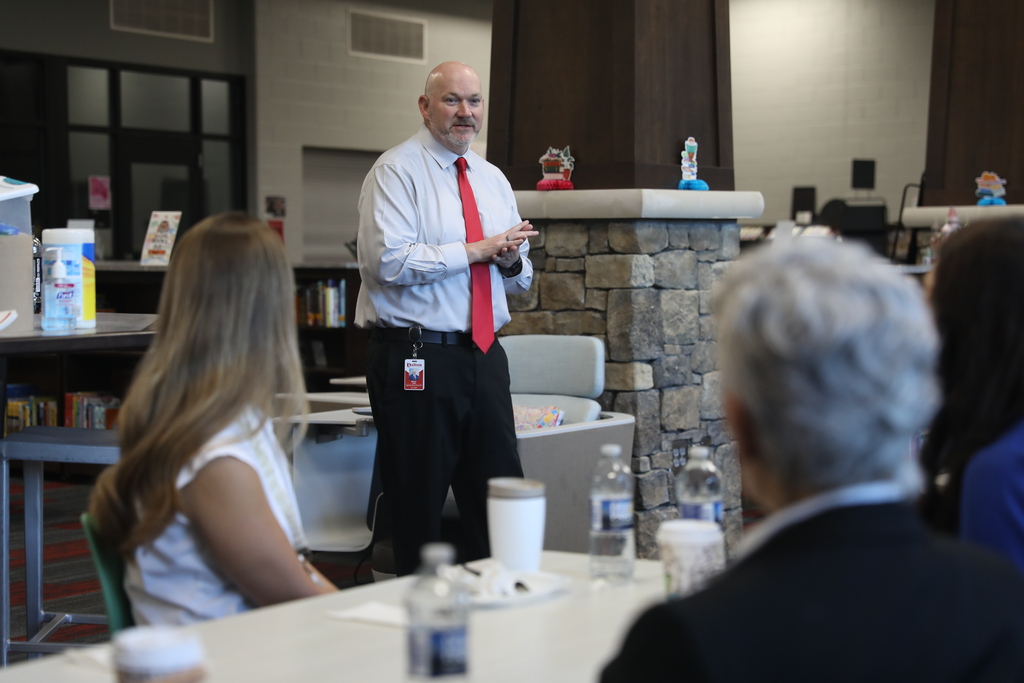A man wearing a white dress shirt and bright red tie speaks to a seated group during a meeting or presentation in a modern library or media center. He stands near a stone column with his hands clasped while attendees sit around tables in the foreground listening. Water bottles, coffee cups, and supplies are visible on the tables, creating a professional workshop atmosphere.