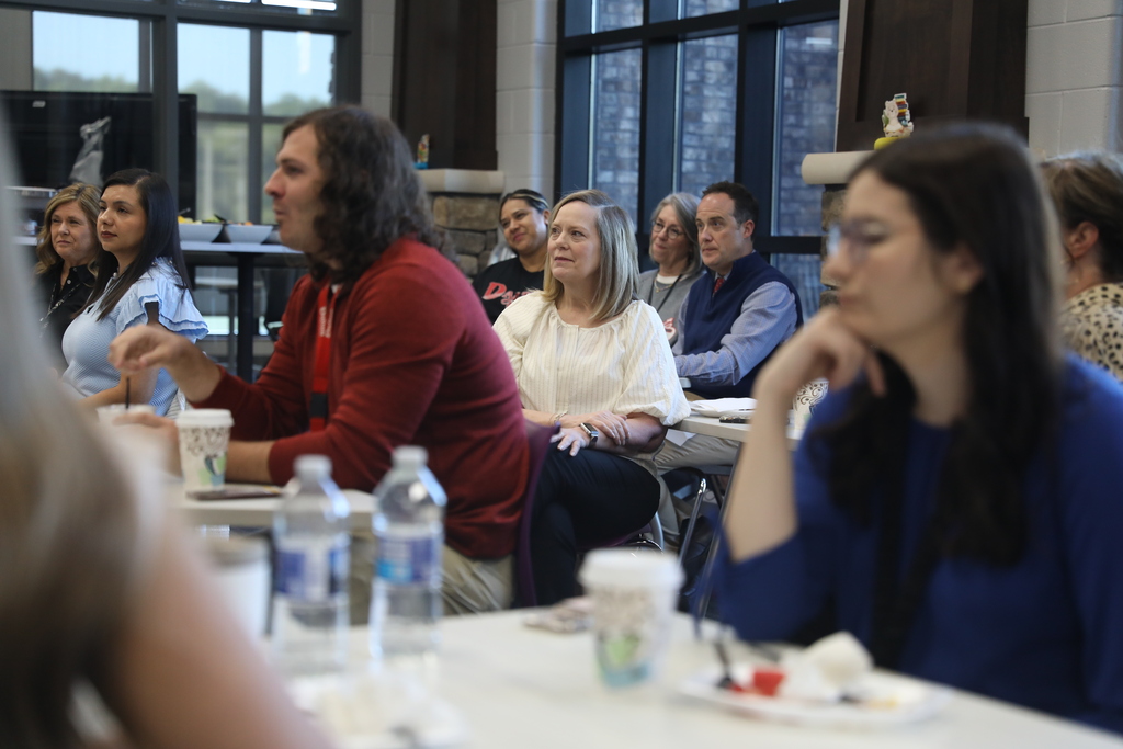 A group of educators and staff members sit around tables during a meeting or professional development session in a bright library or media center. Several attendees are focused on a speaker out of frame, while coffee cups, water bottles, and plates are scattered across the tables. Large windows and stone columns create a warm, collaborative atmosphere.