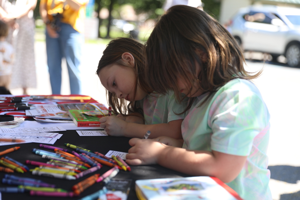 **Alt text:** Two young children sit side by side at an outdoor activity table, concentrating as they color and complete literacy-themed activity sheets. Colorful crayons are scattered across the table alongside books, bookmarks, and educational materials. Sunlight illuminates the scene while the children focus on their work, highlighting a hands-on learning experience that encourages creativity, reading, and community engagement.