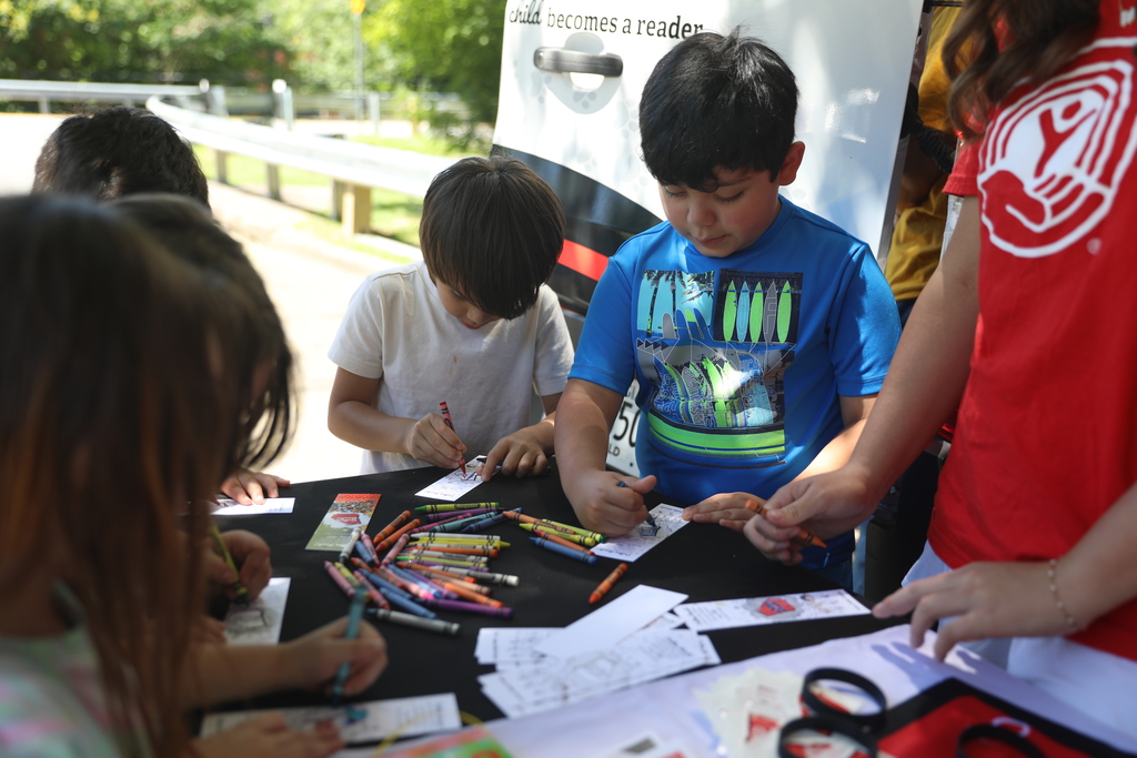 Alt text: Several young children gather around an activity table outdoors, coloring and drawing on small worksheets with crayons. A child in a blue shirt focuses on coloring while another child nearby writes with a crayon. The table is covered with colorful crayons, activity sheets, and bookmarks. In the background, part of a mobile literacy vehicle is visible, suggesting a reading or community literacy event. The image captures students participating in a hands-on educational activity that encourages creativity and engagement with reading.