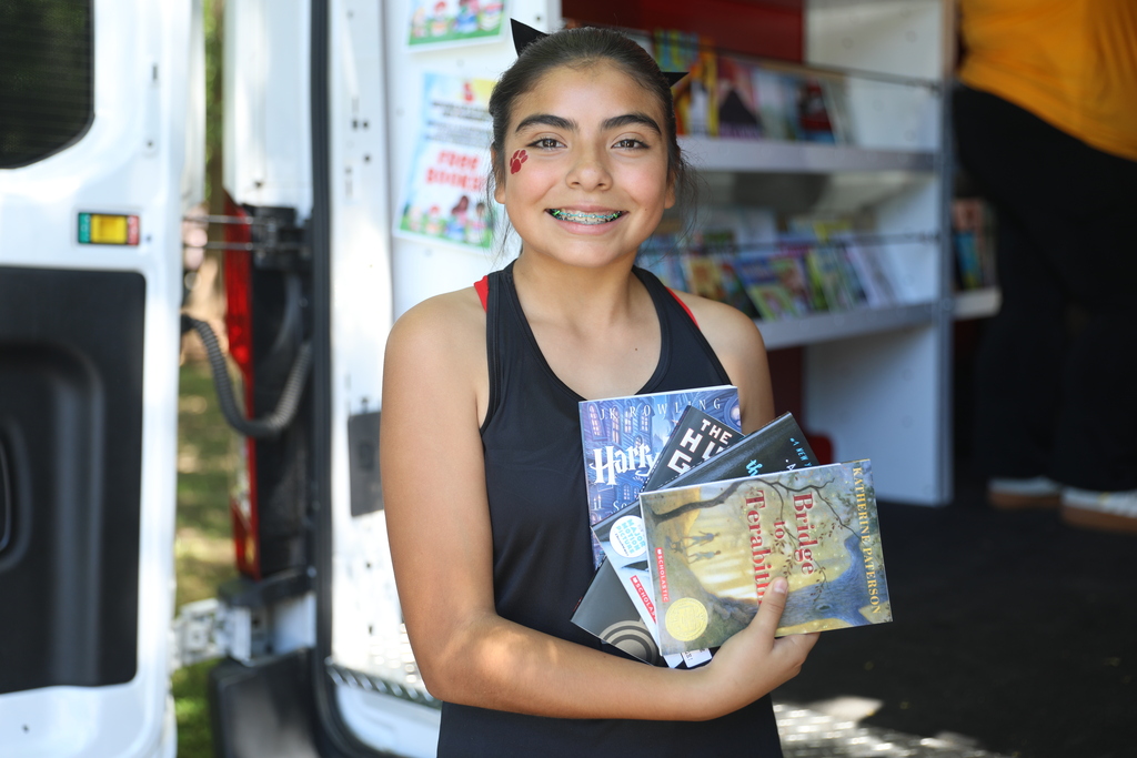 A smiling student stands at the open door of a mobile library, holding a stack of books in her arms. She wears a black athletic top and has a small paw-print face paint design on her cheek. The books she is carrying include popular youth titles, and shelves filled with additional books are visible inside the mobile library behind her. The image highlights student engagement with reading and access to books through a school literacy outreach program.