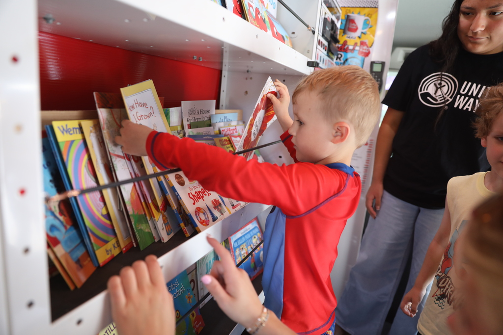 **Alt text:** A young student browses books inside a mobile library, reaching toward a shelf filled with colorful children’s titles. The child, dressed in a red-and-blue outfit, carefully examines a book while other children and an adult stand nearby. Brightly illustrated picture books line the shelves, creating an inviting reading environment. The image captures students exploring book choices and engaging with literacy opportunities through the mobile library program.