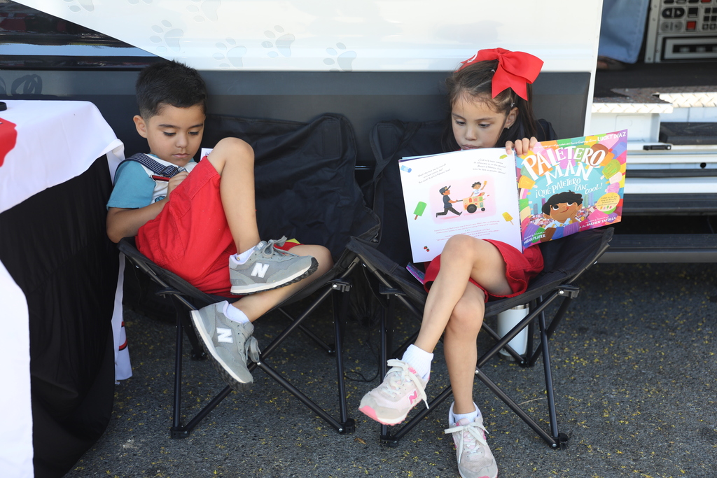 **Alt text:** Two young children sit in folding camp chairs beside a mobile literacy bus, quietly enjoying books during a community reading event. One child reads independently while the other is engaged with a colorful picture book titled *Paletero Man*. Dressed in bright summer clothing, the children appear focused and comfortable, illustrating a welcoming environment that encourages reading, learning, and a love of books.