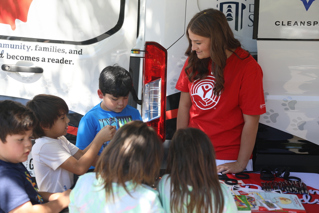**Alt text:** Several young children gather around an activity table at an outdoor literacy event while a student volunteer in a red United Way T-shirt smiles and interacts with them. One child holds up a completed activity while another looks down at their work. The group stands beside a Dalton Public Schools literacy vehicle, which is visible in the background. Activity sheets, bookmarks, and educational materials are spread across the table, creating an engaging environment that encourages reading, learning, and community involvement.