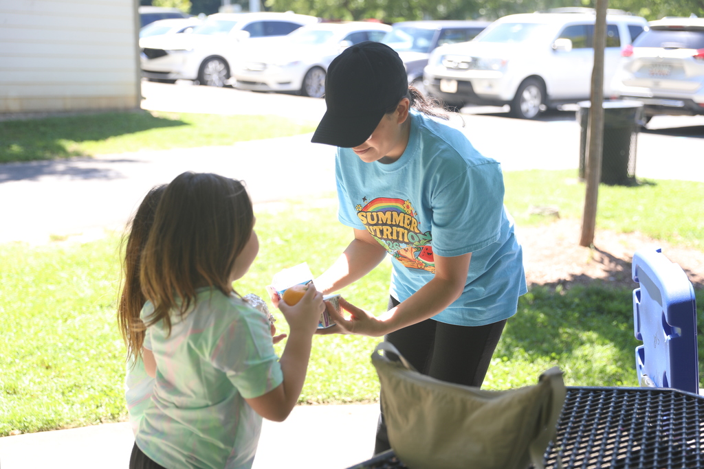 **Alt text:** A staff member wearing a “Summer Nutrition” T-shirt hands food items to a young child during an outdoor summer meal program. The child stands beside a picnic table while receiving lunch, which includes fruit and packaged items. Bright sunlight and a grassy park setting create a welcoming atmosphere, highlighting efforts to provide nutritious meals and support children and families during the summer months.