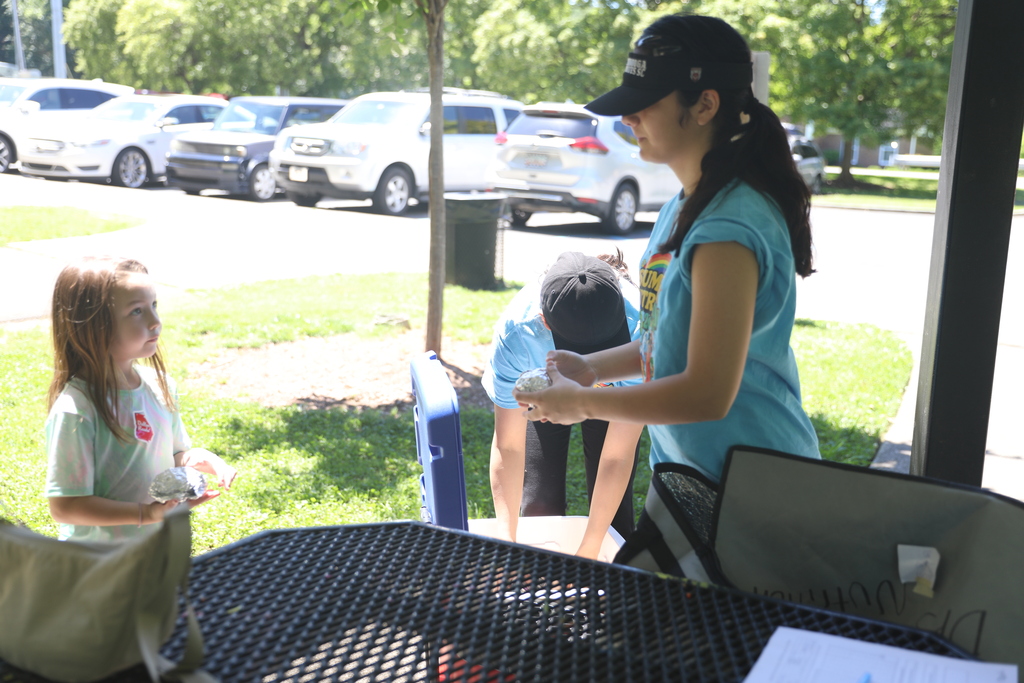 **Alt text:** A child participates in a summer meal program while staff members distribute food and assist families at an outdoor park pavilion. One staff member wearing a “Summer Nutrition” T-shirt prepares meal items from a cooler while another helps nearby. The child holds a wrapped meal and looks toward the volunteers. Parked vehicles and green space in the background create a community-centered setting that highlights access to nutritious meals and supportive services for children during the summer months.