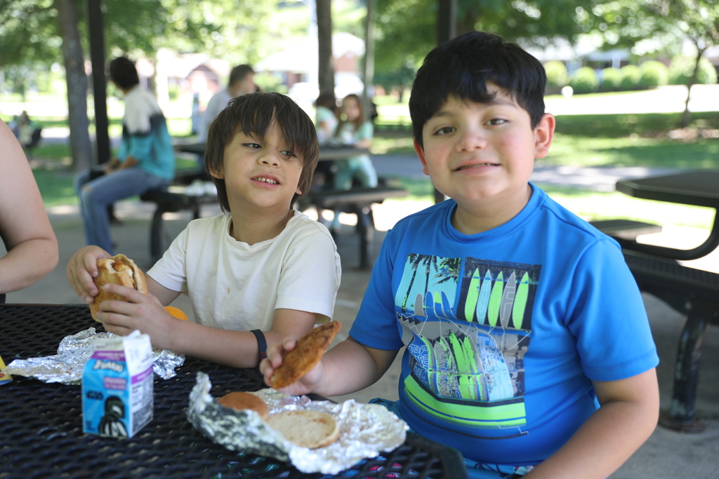 **Alt text:** Two young children sit together at a picnic table in a shaded park pavilion, enjoying lunch during a community event. One child holds a sandwich while the other smiles toward the camera and holds a slice of pizza. A milk carton, wrapped food items, and fruit rest on the table in front of them. Other families can be seen in the background, creating a welcoming atmosphere that highlights community connection, family engagement, and shared experiences.