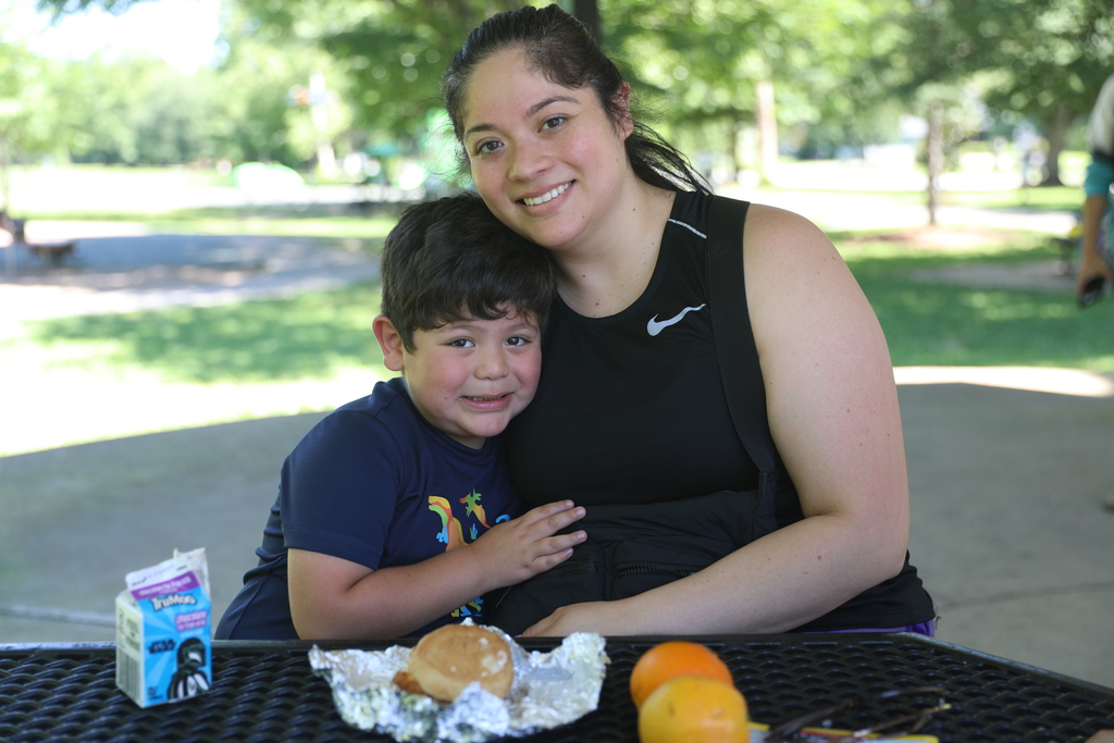 A young child and an adult sit together at a picnic table in a shaded park pavilion, smiling toward the camera. The child leans comfortably against the adult while enjoying lunch. On the table are a milk carton, a sandwich wrapped in foil, and pieces of fruit. The scene conveys a warm moment of connection and family engagement during a community event, with trees and green space visible in the background.