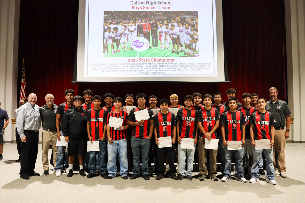 Alt text: Group photo of the Dalton High School boys soccer team standing on a stage with coaches and school leaders during a recognition ceremony. Team members wear black-and-red Dalton soccer jerseys, and several students hold certificates. A large projection screen behind them displays a team photo and recognizes the Dalton High School Boys Soccer Team as the 2026 State Champions. Red theater curtains frame the stage, and the group poses together to celebrate their championship season and achievements.