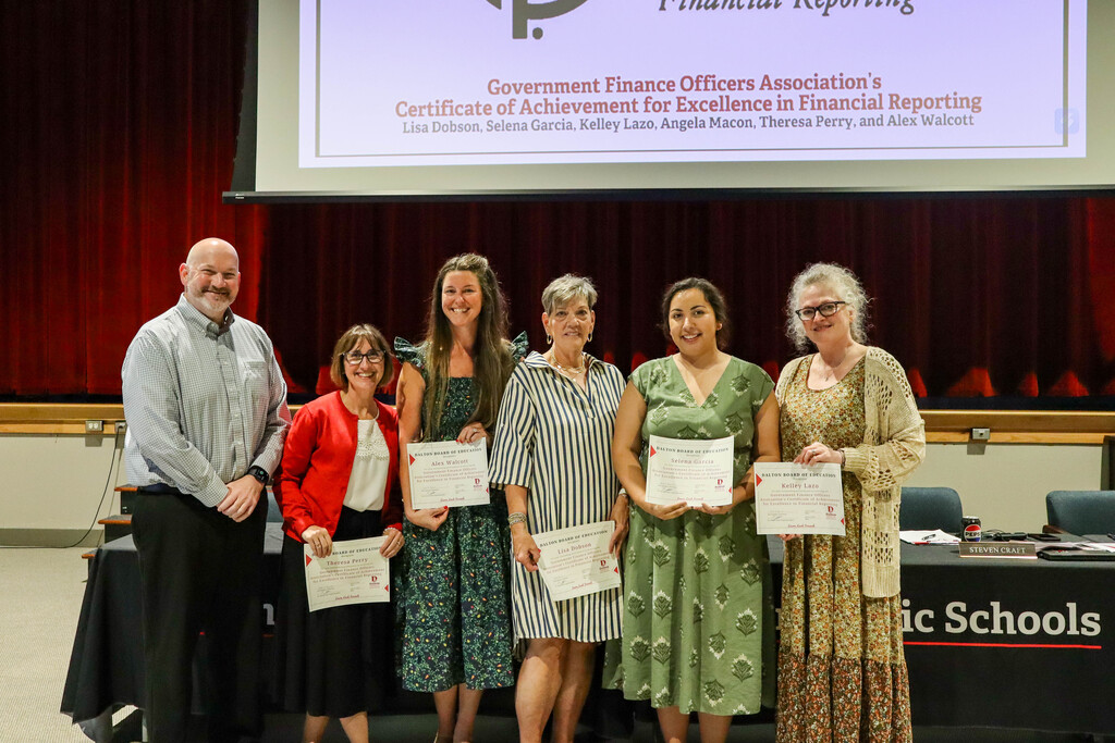 **Alt text:** Group photo of Dalton Public Schools staff members being recognized during a Board of Education meeting for excellence in financial reporting. Six individuals stand together in front of a stage with red curtains, several holding certificates. A presentation screen above them highlights the Government Finance Officers Associationās Certificate of Achievement for Excellence in Financial Reporting and lists the recognized team members. Meeting tables with microphones and Dalton Public Schools branding are visible in the foreground, marking the districtās recognition of the finance departmentās accomplishment.