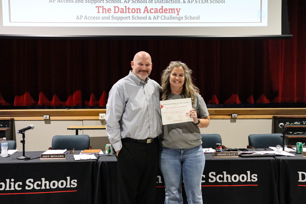 **Alt text:** Two adults pose together in front of a stage during a Dalton Public Schools Board of Education meeting. One person holds a certificate recognizing The Dalton Academy as an **AP Access and Support School** and **AP Challenge School**. Behind them are meeting tables with microphones, nameplates, and Dalton Public Schools branding. A large projection screen above the stage displays information about The Dalton Academyās Advanced Placement recognitions. The photo captures the presentation of the schoolās achievement and recognition for expanding access to rigorous academic opportunities.