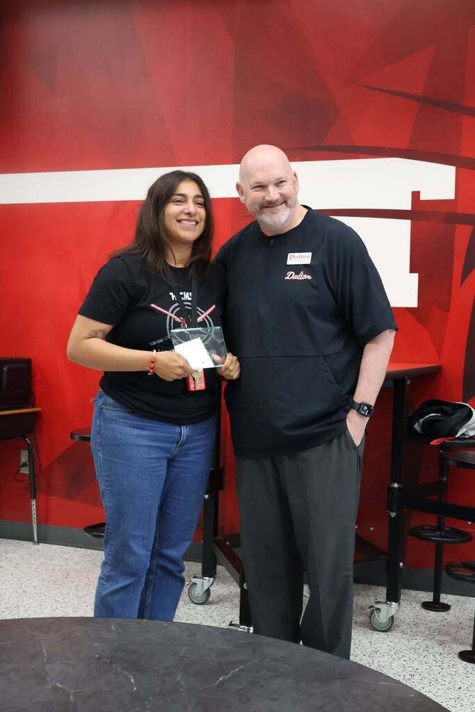 Two people pose together indoors in front of a red-and-white Dalton-themed wall. One person holds a glass award while smiling for the camera, and the other stands beside them wearing a black Dalton-branded pullover and name badge. The setting appears to be a school gathering or employee recognition event, with cafeteria-style tables and stools visible in the background. The photo captures a moment of appreciation and recognition within the Dalton Public Schools community.