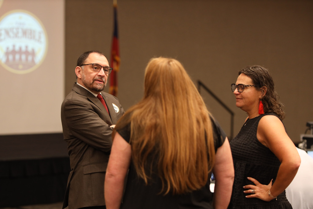**Alt text:** Three Dalton Public Schools staff members stand together in conversation during a leadership event. One staff member in a suit and red tie speaks while two others listen nearby. In the background, part of a presentation screen displaying the "Ensemble" leadership archetype is visible, along with the stage and a Georgia state flag, highlighting a moment of collaboration and professional dialogue.