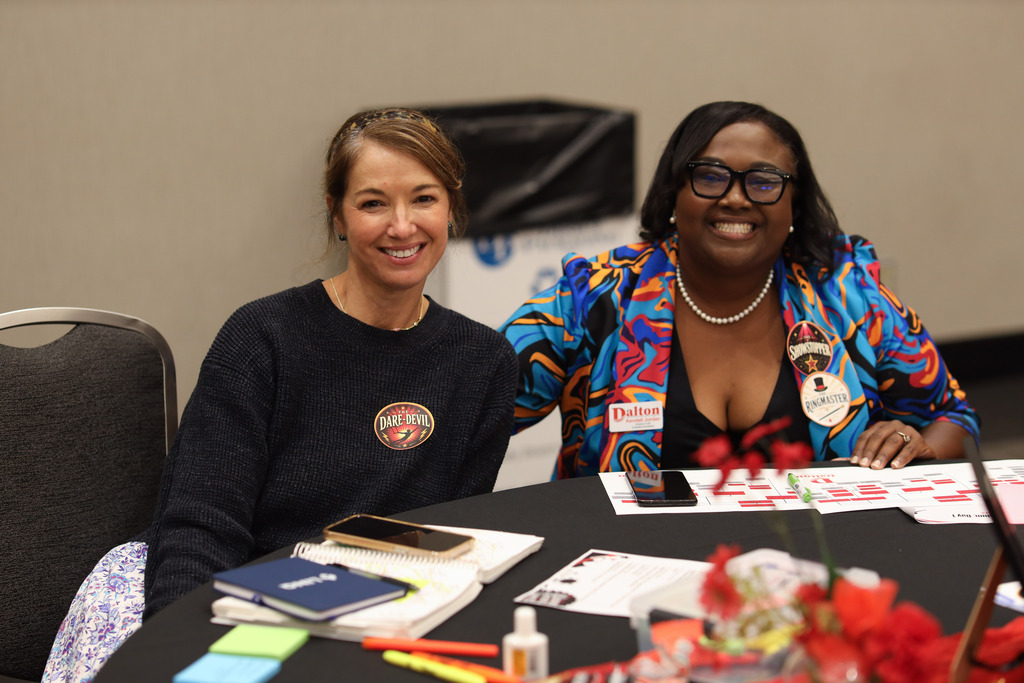 **Alt text:** Two Dalton Public Schools staff members smile at the camera while seated together at a table during a district professional learning event. One participant wears a dark sweater with a "Daredevil" sticker, while the other wears a colorful patterned blazer with themed "Ringmaster" and "Showstopper" stickers. The table is covered with workshop materials, including notebooks, sticky notes, markers, a phone, and printed activity sheets. A small floral centerpiece adds color to the table, reflecting the collaborative and engaging atmosphere of the event.