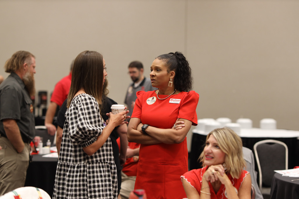 **Alt text:** Several Dalton Public Schools staff members visit and build connections during a district professional learning event. At the center, two staff members are engaged in conversation, one holding a coffee cup while the other, wearing a bright red dress with a themed "Ringmaster" sticker, listens attentively with her arms crossed. Other participants are chatting and seated around the room in the background, creating a welcoming and collaborative atmosphere before or between sessions.