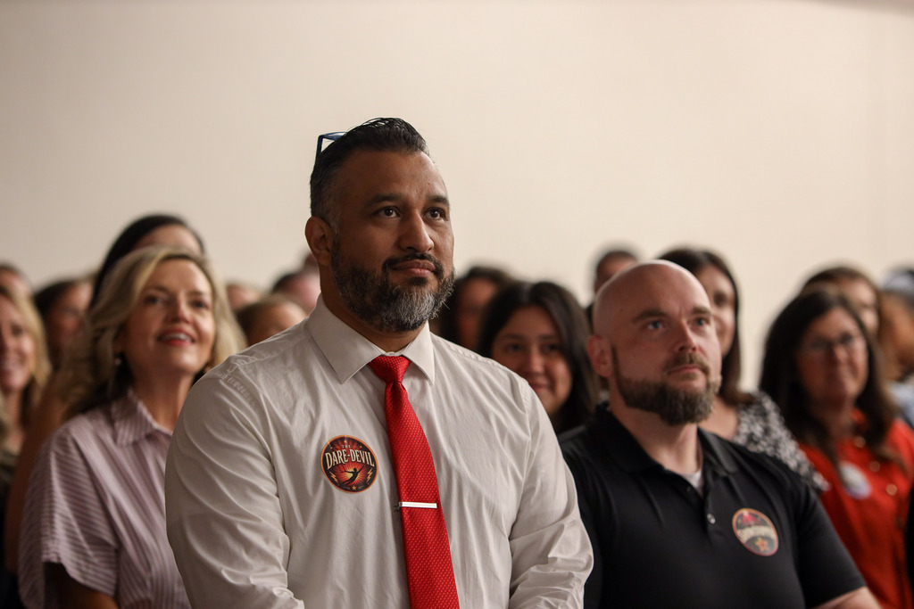 A man wearing a white dress shirt, red tie, and a round "Daredevil" sticker stands attentively in an audience during a Dalton Public Schools event. He has a salt-and-pepper beard and glasses resting on top of his head. Around him, other staff members are seated and focused on the presentation, including a man in a black polo shirt wearing a matching themed sticker. The room is filled with attendees, creating a warm, engaged atmosphere as participants listen to the speaker.