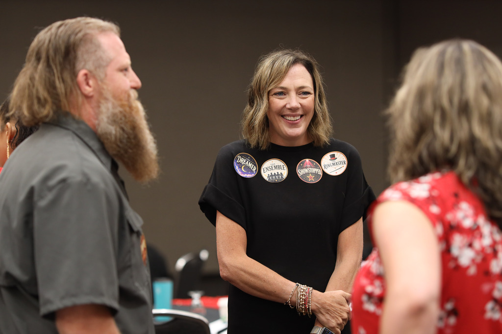 **Alt text:** Three Dalton Public Schools staff members stand together in conversation during a district professional learning event. At the center, a smiling staff member wearing a black dress is engaged with colleagues and displays several themed event stickers, including "Dreamer," "Ensemble," "Showstopper," and "Ringmaster." Two other participants are seen from the side and back as they chat, creating a candid moment of collaboration and connection. The photo highlights relationship-building and teamwork in a welcoming professional learning environment.