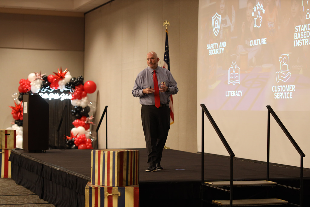 **Alt text:** Superintendent Steven Craft walks across the stage while presenting during a Dalton Public Schools leadership event. A large presentation screen beside him highlights the district's five strategic commitments: Safety and Security, Positive Culture, Standards-Based Tier 1 Instruction, Literacy, and Customer Service, each represented with an icon. The stage features red, black, and white balloon decorations, circus-themed props, and a "Step Right Up" backdrop, reinforcing the event's leadership theme.