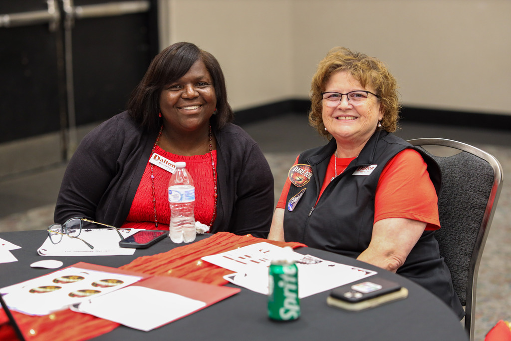 **Alt text:** Two Dalton Public Schools staff members smile at the camera while seated together at a round table during a district professional learning event. One wears a red blouse with a black cardigan, and the other wears a red shirt with a black vest decorated with themed event stickers, including a "Daredevil" sticker. The table is set with event materials, papers, a water bottle, a soft drink, and a phone, reflecting an active workshop setting. The photo captures a welcoming, collaborative atmosphere as participants engage in the day's activities.