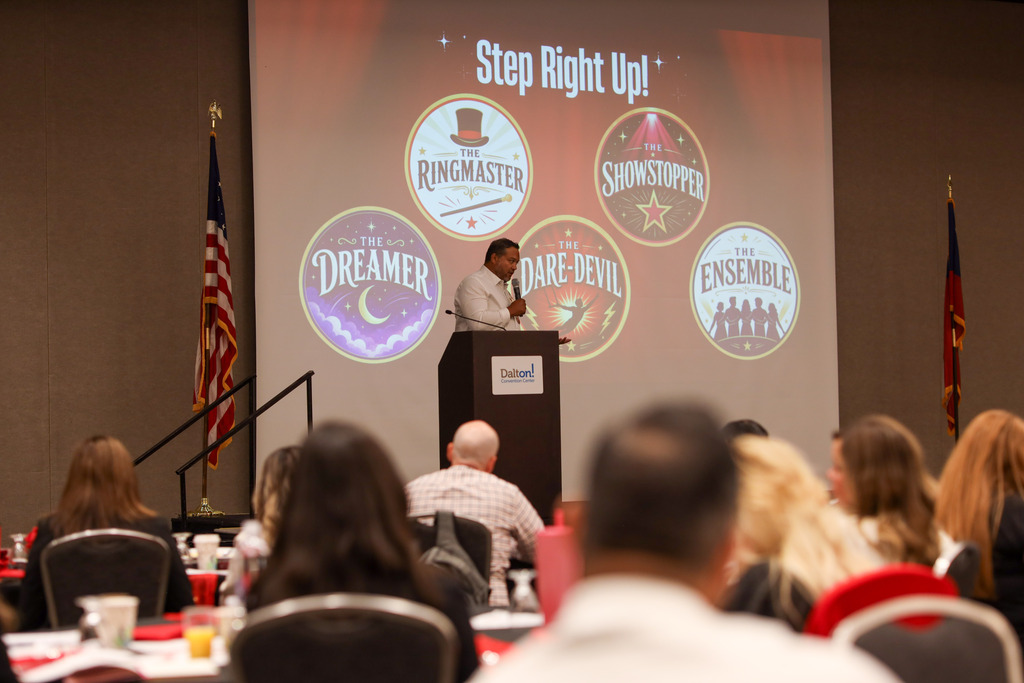 **Alt text:** A presenter stands at a podium speaking to Dalton Public Schools staff during a leadership event at the Dalton Convention Center. Behind the speaker, a large presentation screen displays the words "Step Right Up!" alongside five circus-themed leadership archetypes: The Ringmaster, The Showstopper, The Dreamer, The Dare-Devil, and The Ensemble. Staff members are seated at round tables in the foreground, listening as the presentation introduces the event's interactive activity and theme.