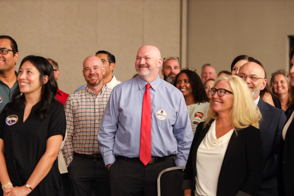 A group of Dalton Public Schools staff members stand together smiling and watching a presentation during a district event. At the center, Superintendent Dr. Steven Craft wears a blue dress shirt, red tie, and a themed "Ringmaster" sticker, smiling as he looks toward the speaker. Around him, colleagues in business and business-casual attire are also smiling and engaged, creating a warm, celebratory atmosphere. The photo captures staff enjoying a shared moment during the event.