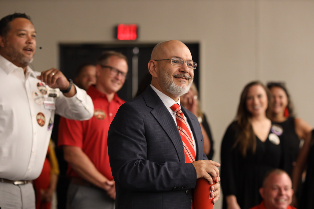 **Alt text:** A Dalton Public Schools administrator in a navy suit and red striped tie smiles while participating in a leadership activity during a district event. Other staff members stand nearby watching and smiling, many wearing themed role stickers that align with the event's leadership activities. The candid moment captures staff engagement and a positive, collaborative atmosphere.