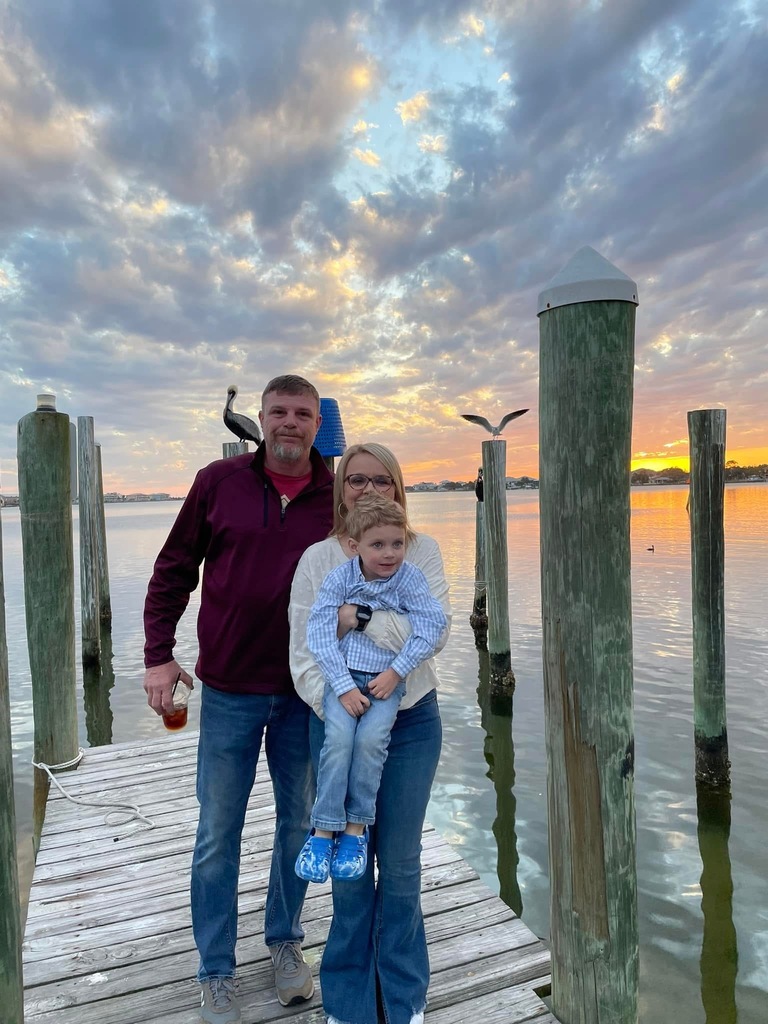 Man, woman, and child stand on dock by the water at sunset.