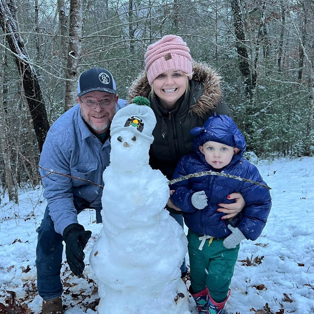 man, woman and child stand next to snowman.