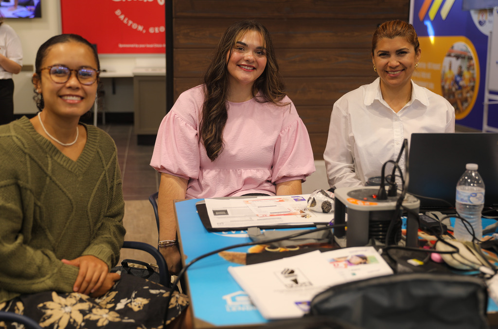Three women seated at a table equipped with power charging hubs and laptops smile at the camera. On the left, a woman wears glasses and an olive green sweater; in the middle, a woman wears a pink puff-sleeve top; on the right, a woman wears a white button-down shirt.