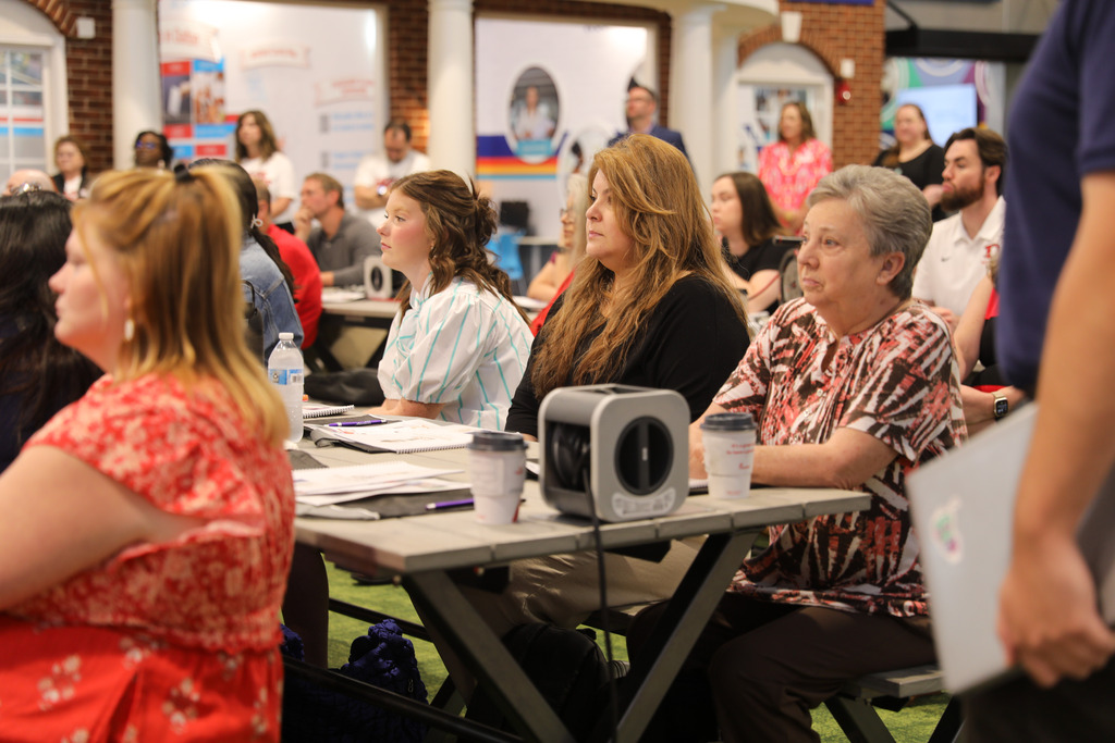 Profile view of audience members seated at a long gray table during a presentation. In focus near the middle, a woman with long light brown hair in a black top listens intently beside an older woman with short gray hair in a patterned blouse.