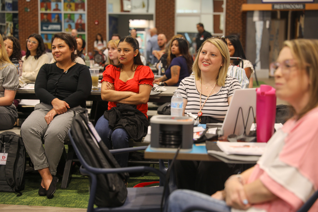 Attendees seated at workshop tables watching a speaker off-camera. In the middle ground, a blonde woman in a black-and-white striped shirt smiles while looking toward the stage area.
