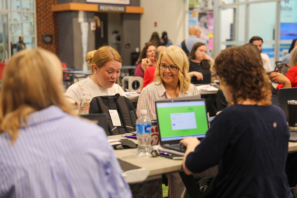Attendees seated at a long table during a workshop. In the center, a blonde woman with glasses wearing a striped button-down shirt smiles while listening, as a colleague in a navy top works on a laptop in the foreground.