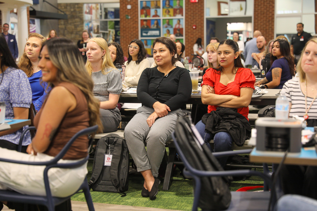 Event attendees seated on benches along a table, listening attentively to a presenter off-camera. In the center, a woman in a black cardigan and houndstooth-patterned pants rests her clasped hands in her lap beside a colleague in a bright red top with crossed arms.