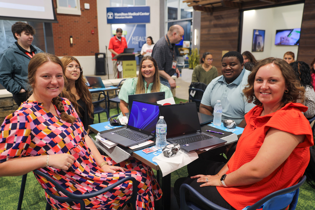 Five event participants sit around a square table with open laptops and water bottles, smiling toward the camera. In the foreground, a woman on the left wears a brightly colored geometric patterned dress, while a woman on the right wears a bright red top.