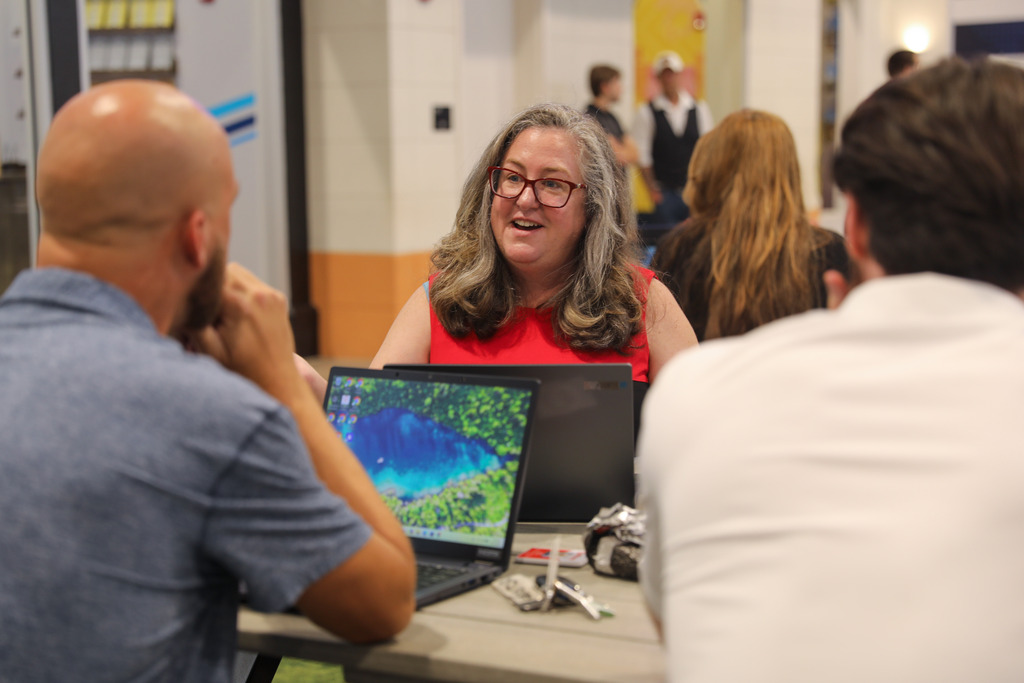 A woman with long wavy graying hair, red-framed glasses, and a red sleeveless top sits at a table speaking warmly to two male colleagues. An open laptop displaying a scenic landscape wallpaper sits on the table between them.
