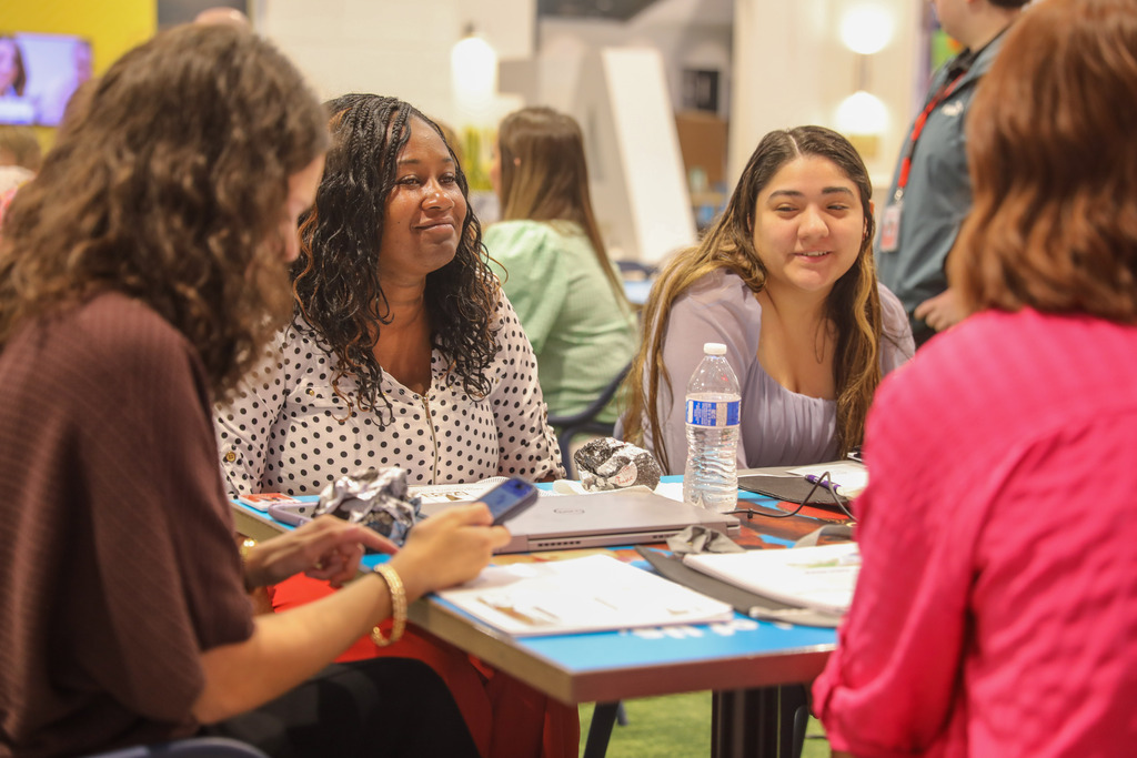 A group of women sitting at a blue-topped table during a workshop. A woman with dark curly hair in a white polka-dot top smiles while listening to a speaker across the table, with laptops, water bottles, and notebooks spread out before them.