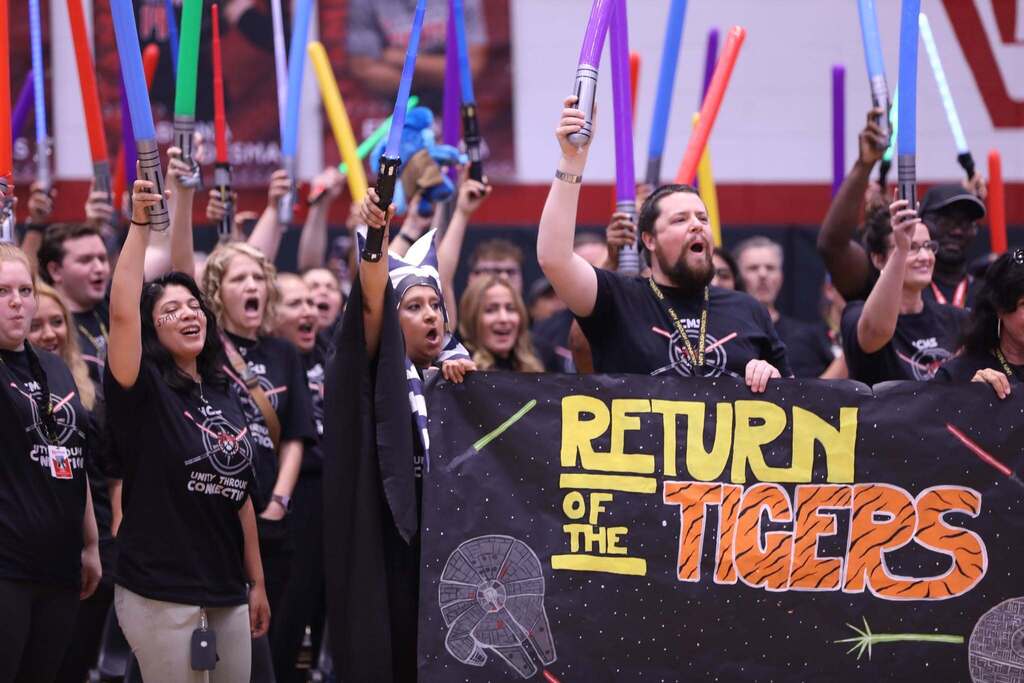 A lively group of teachers and staff holding colorful lightsabers high in the air inside a gymnasium. In the center, two people hold a large black banner that reads "RETURN OF THE TIGERS" in bold Star Wars-style yellow and tiger-striped lettering, complete with drawings of the Millennium Falcon and the Death Star. One staff member is wearing an Ahsoka Tano headpiece, and several others are wearing matching black "Unity Through Connection" t-shirts. 