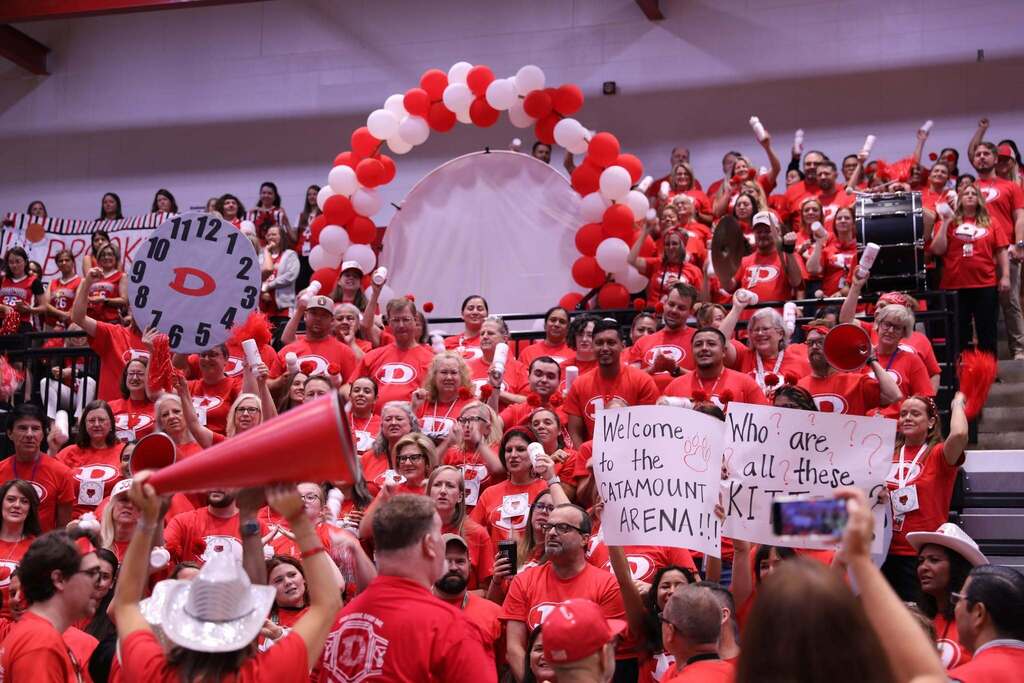 A crowd of school staff wearing red t-shirts gathered in gym bleachers under a large arch of red and white balloons. Many people are cheering, holding red pom-poms, and holding signs, including one that reads "Welcome to the CATAMOUNT ARENA!!!" and another holding a giant cardboard clock set to 12:00 with a red letter 'D' in the center. 