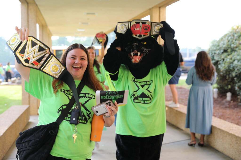 A woman smiling brightly and a school mascot in a black wildcat costume walking down a covered outdoor walkway. Both are wearing bright neon green t-shirts and triumphantly holding up replica WWE championship belt trophies. The woman holds her belt up to the camera while the mascot balances another belt on top of its head. 