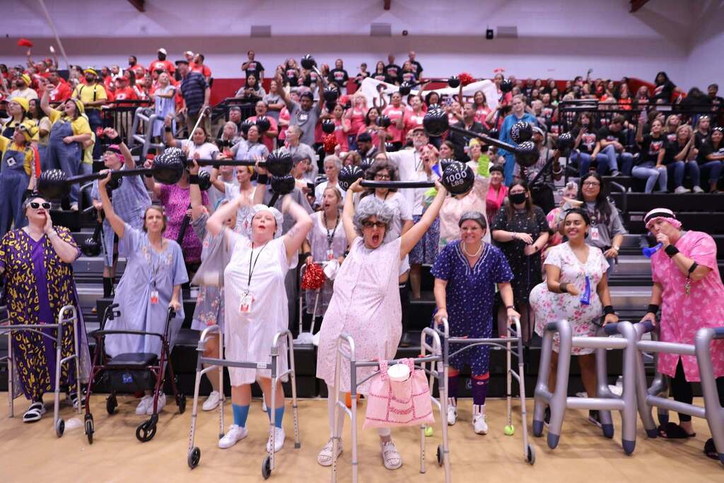 A high-energy group of staff members in gym bleachers dressed as grandmothers in floral nightgowns, gray wigs, and hair curlers. Several hold up medical walkers and fake, oversized prop barbells labeled "1000 LBS" while cheering and making excited faces. 