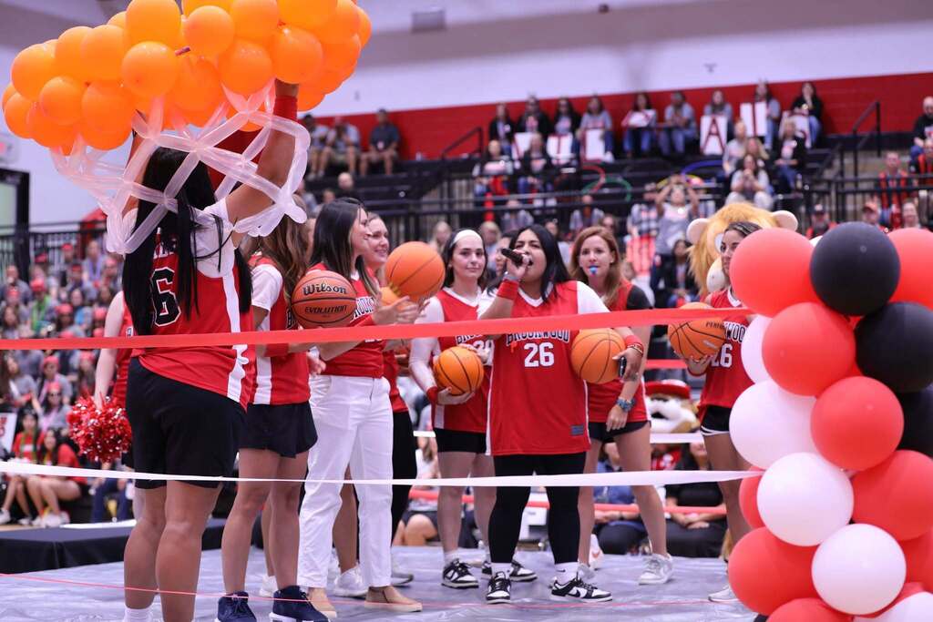 A group of school staff members in red Brookwood jerseys and white pants or shorts standing in a gym during an event. One woman speaks into a microphone, surrounded by colleagues holding basketballs. On the left, someone holds up a large cluster of bright orange balloons wrapped in white netting, and a red and white ribbon stretches across the foreground. 