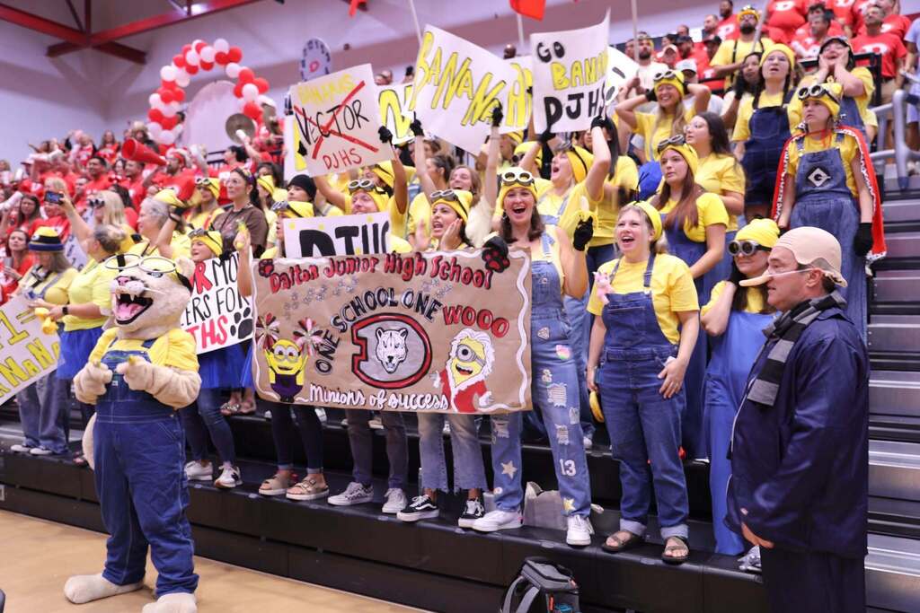 An energetic crowd of school staff seated in bleachers dressed in Despicable Me Minion-themed costumes, wearing yellow shirts, denim overalls, and yellow beanies with Minion goggles. One person is dressed as Gru in a blue jacket and striped scarf with a prosthetic nose. In the center, staff members hold up signs and a large painted banner that reads "Dalton Junior High School - ONE SCHOOL ONE WOOO - Minions of Success," flanked by a mascot in a Minion costume. 