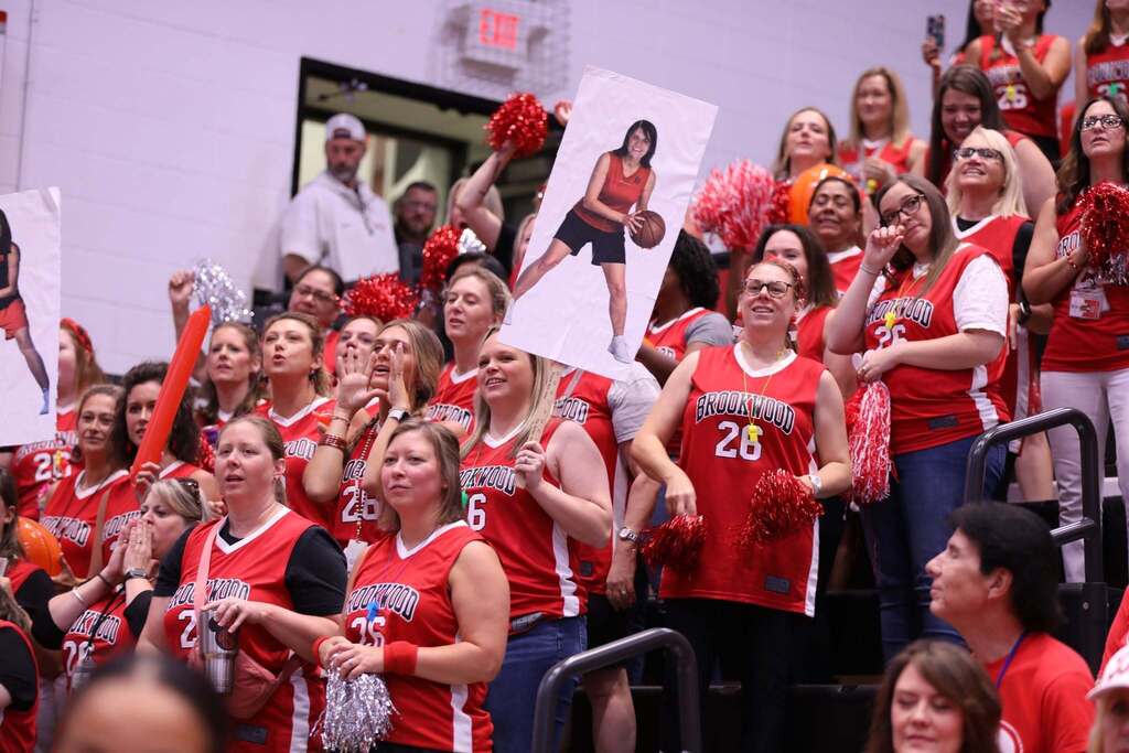 A crowd of energetic school staff in bleachers wearing red "Brookwood" basketball jerseys. Many are holding red and silver pom-poms, while a woman near the center holds up a tall poster showing a photo of a staff member posing with a basketball. 