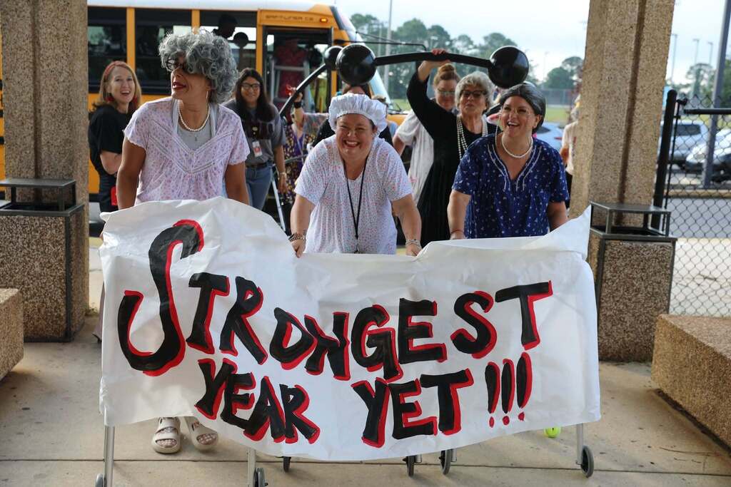 Three women dressed as grandmothers with gray wigs, pearl necklaces, and floral nightgowns walking outdoors under a covered walkway. They laugh while holding up a large hand-painted banner that reads "STRONGEST YEAR YET!!!" attached to their rolling walkers, as another person holds a prop barbell overhead behind them. 