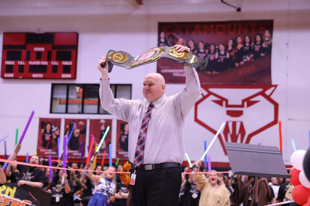 A man in a light gray dress shirt and tie standing indoors, proudly holding a large championship belt trophy high above his head with both hands. In the background, audience members cheer while holding up glowing, colorful lightsabers. 